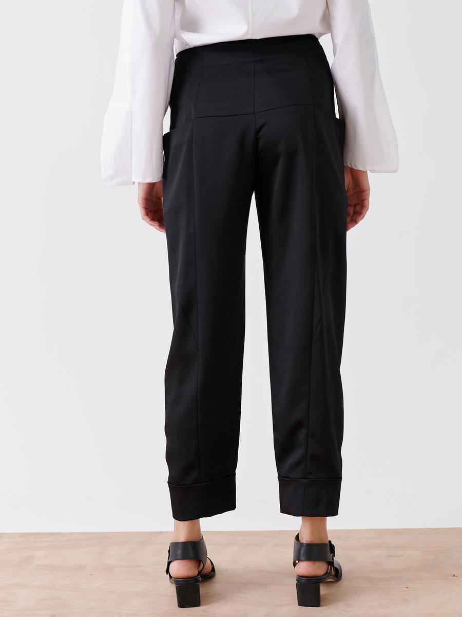 Rear view of a person wearing black tailored pants from the Akeo Jodhpur - Final Sale by Zero + Maria Cornejo, styled with a white blouse featuring flared sleeves and black heeled sandals, standing on a wooden floor against a plain white background.