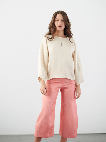 A woman with long hair, wearing a beige blouse and Zero + Maria Cornejo's Curved Eda Culotte in pink eco linen, poses against a white background with a neutral expression, embodying the elegance of sustainable fashion.