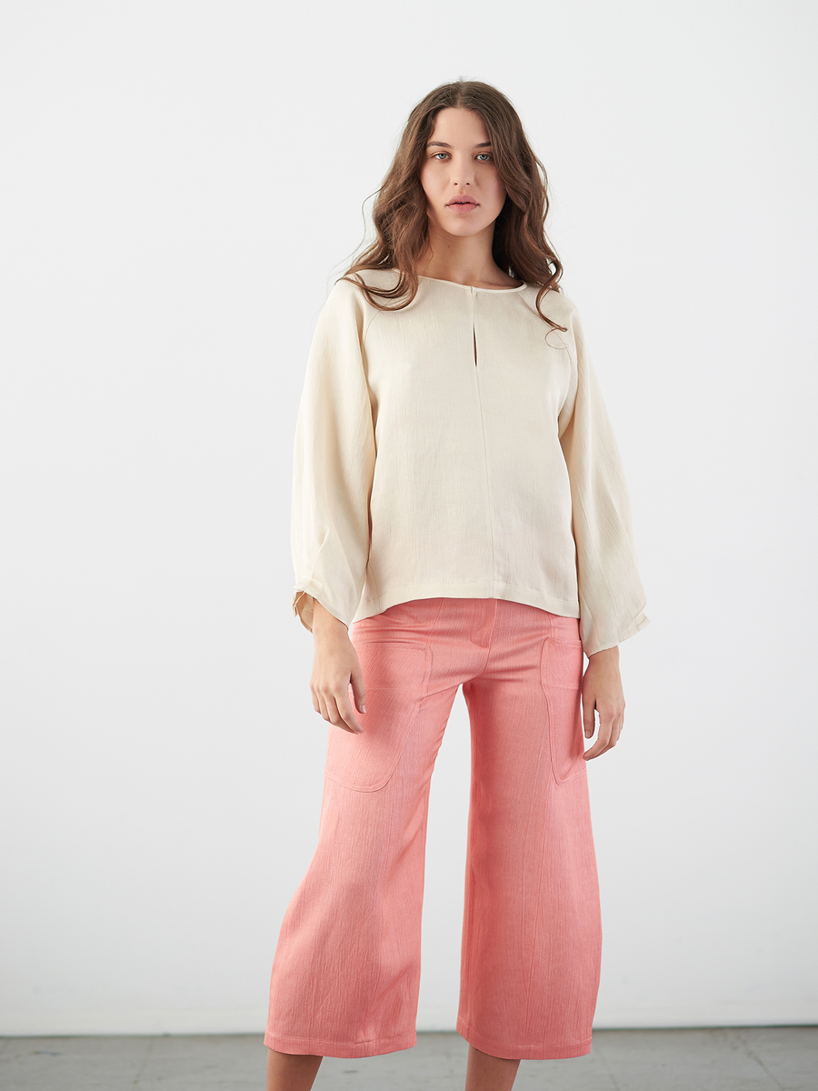 A woman with long hair, wearing a beige blouse and Zero + Maria Cornejo's Curved Eda Culotte in pink eco linen, poses against a white background with a neutral expression, embodying the elegance of sustainable fashion.