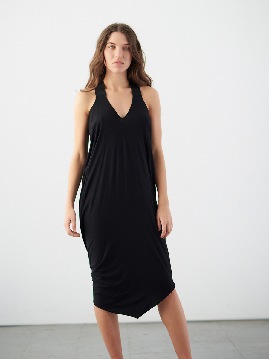 A person with long hair, wearing the Long Ibit Dress in a sustainable black V-neck midi style by Zero + Maria Cornejo, stands against a white background, showing a neutral expression and hands resting by their sides.