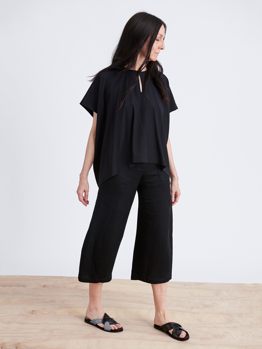 A woman with long hair stands on a wooden floor, wearing an eco-friendly loose black top and Curved Eda Culotte by Zero + Maria Cornejo. She pairs the look with sandals and gazes to her right against a plain white background.