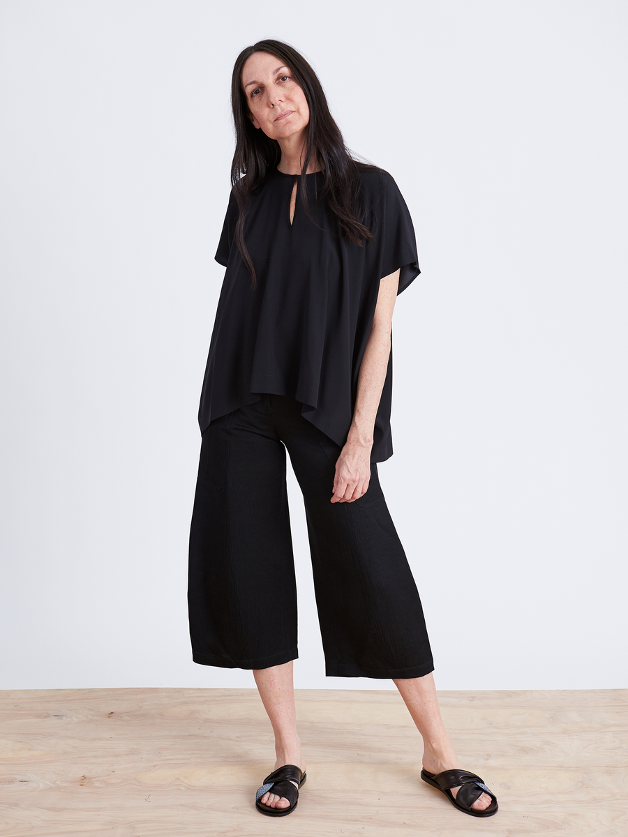 A woman with long dark hair stands against a plain background, wearing a loose black top and the Zero + Maria Cornejo Curved Eda Culotte. She looks at the camera with a relaxed posture, her right hand resting near her hip. Black sandals complete her effortlessly chic look.