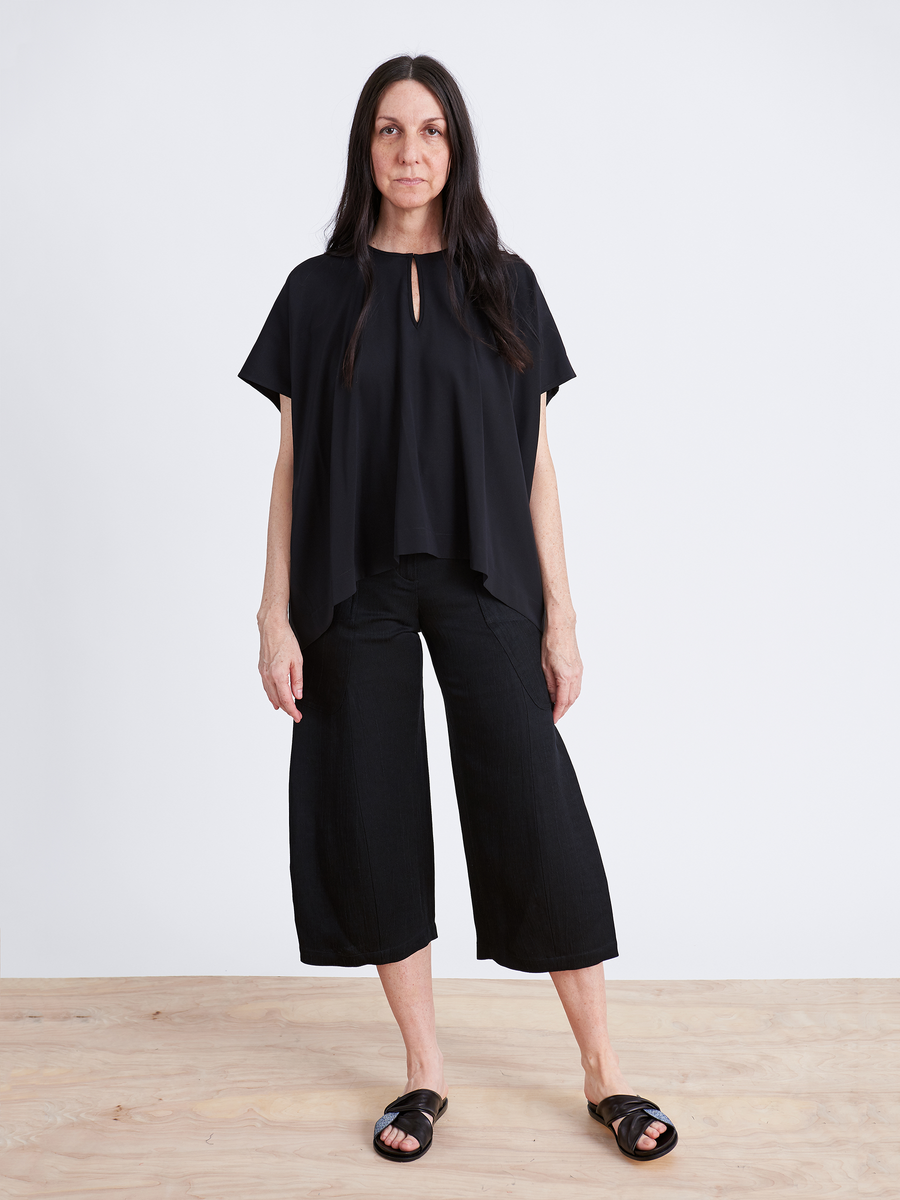 A person with long dark hair stands against a white background wearing a loose black top and the Curved Eda Culotte by Zero + Maria Cornejo, paired with black slide sandals. The flooring is light wood.