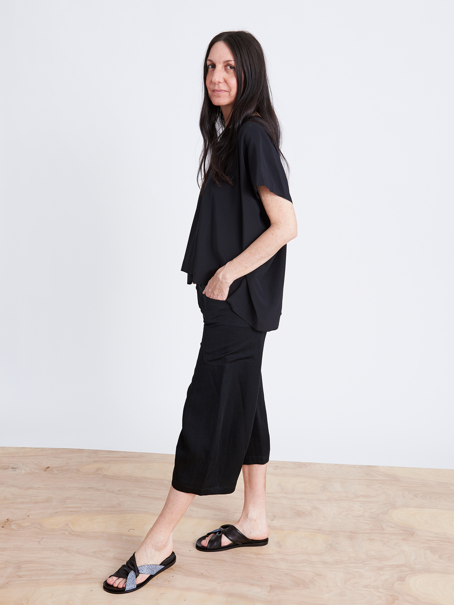 A woman with long dark hair in a black outfit featuring the Curved Eda Culotte by Zero + Maria Cornejo stands on a light wooden floor against a white backdrop, leaning slightly to one side with a relaxed expression, embodying the elegance of sustainable fashion choices.