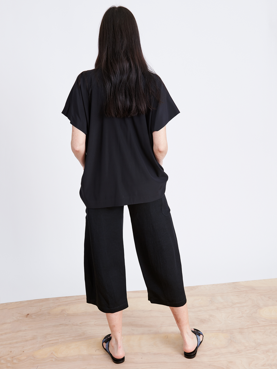 A person with long dark hair stands facing away, wearing a loose black shirt, Curved Eda Culotte by Zero + Maria Cornejo, and black sandals. They are on a light wooden floor against a plain white background, embodying sustainable simplicity.