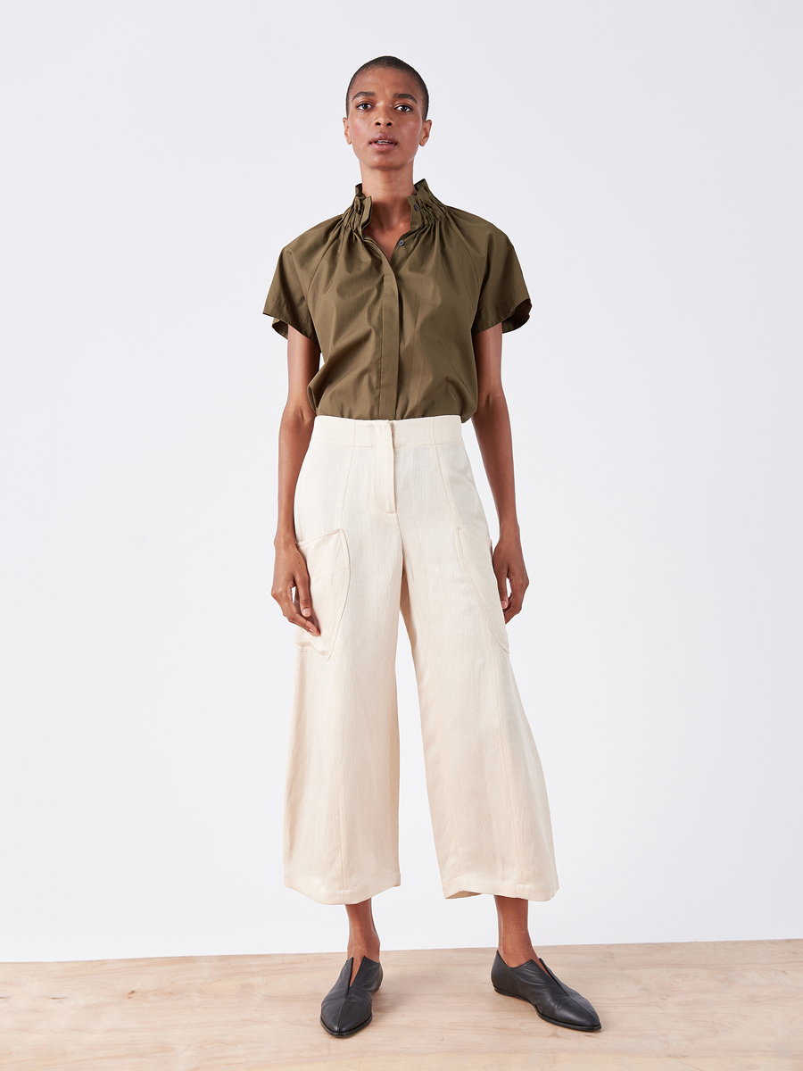 The person poses against a plain white background, wearing an olive green top and the Zero + Maria Cornejo Curved Eda Culotte in eco linen cream. Black slip-on shoes complete this stylish, sustainable look.