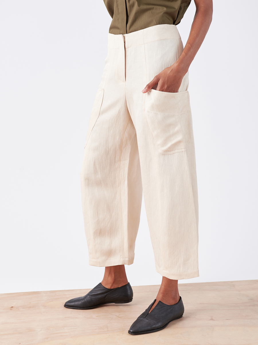 A person stands on a wooden floor, wearing Zero + Maria Cornejo's Curved Eda Culotte in eco-linen cream. The wide-leg, ankle-length pants with large pockets are stylishly paired with a green top and black slip-on shoes for an effortlessly chic look.