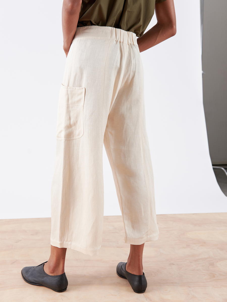 Somebody sports the Zero + Maria Cornejo's Curved Eda Culotte in cream eco linen with a left-side pocket, paired with black slip-on shoes and an olive green short-sleeve top against a minimal, neutral backdrop, emphasizing the sustainable style.