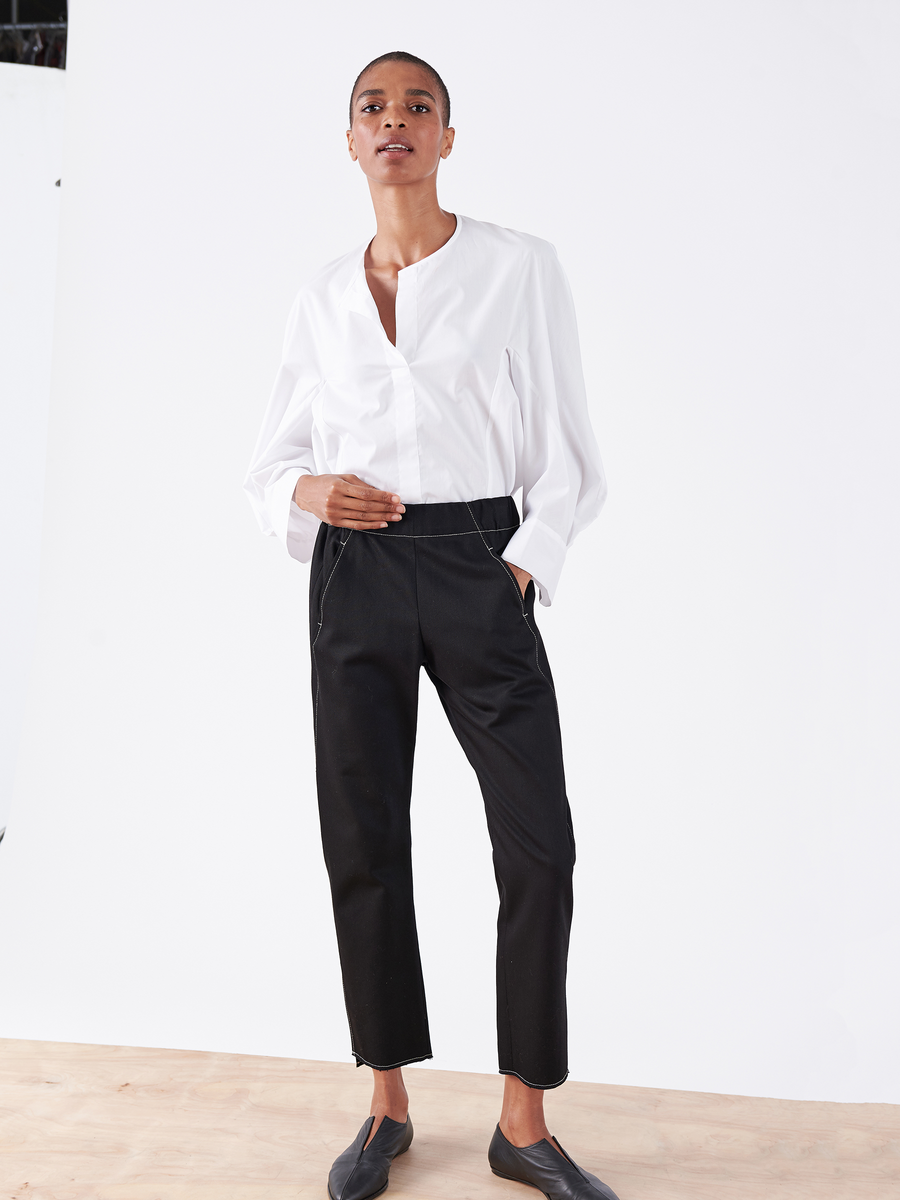 A person stands confidently against a white background wearing a white long-sleeve shirt, black slip-on shoes, and the sustainable Denim Elli Pant by Zero + Maria Cornejo, adding eco-friendly style to their look.