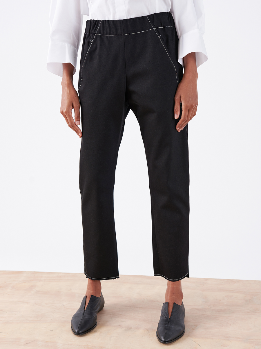 A person is wearing the Denim Elli Pant by Zero + Maria Cornejo, in black eco denim with white stitching, front pockets, and made from 100% organic cotton. They have paired it with black slip-on shoes and a crisp white shirt while standing on a light wooden floor.