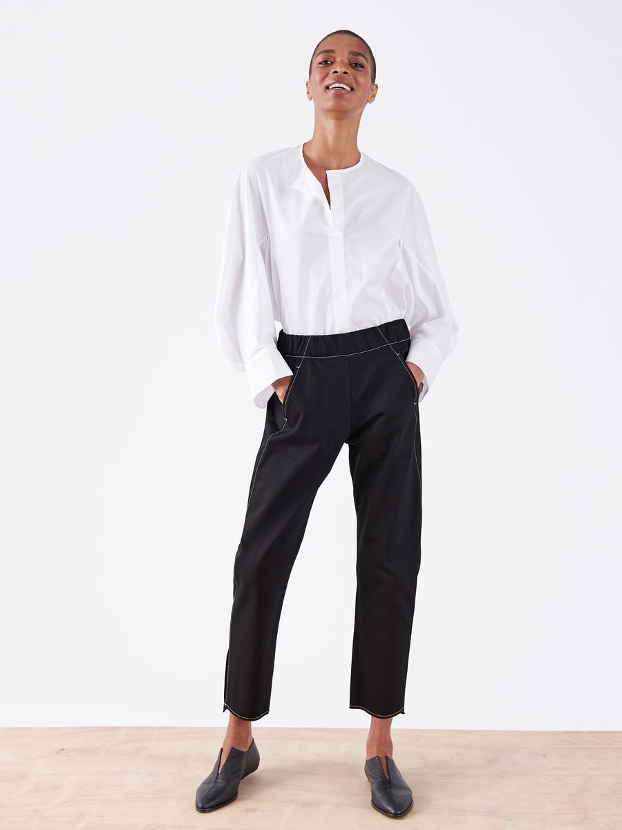 A person stands confidently, smiling in a white long-sleeve shirt and sleek Denim Elli Pant by Zero + Maria Cornejo. Their hands are in their pockets, effortlessly paired with black shoes against a plain white background.