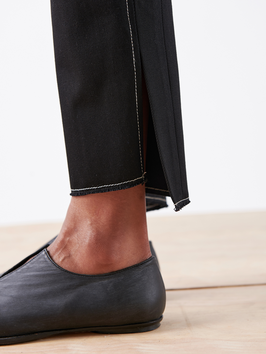 Close-up of a person's lower leg wearing Zero + Maria Cornejo's Denim Elli Pant - Final Sale with a slit at the hem, paired with black slip-on shoes on a light wood floor.