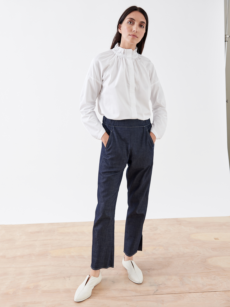 A person with long dark hair stands on a wooden floor wearing a white blouse with a ruffled collar and the Denim Elli Pant by Zero + Maria Cornejo. With hands in pockets, they glance slightly to the side, effortlessly pairing the look with white slip-on shoes.