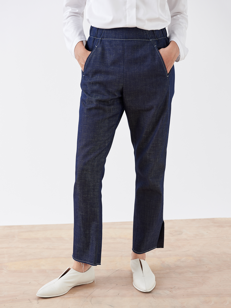 A person wears Zero + Maria Cornejo's Denim Elli Pant in dark blue, featuring a straight cut and subtle slits at the hem, paired with a white shirt and shoes. The background is plain and light-colored.