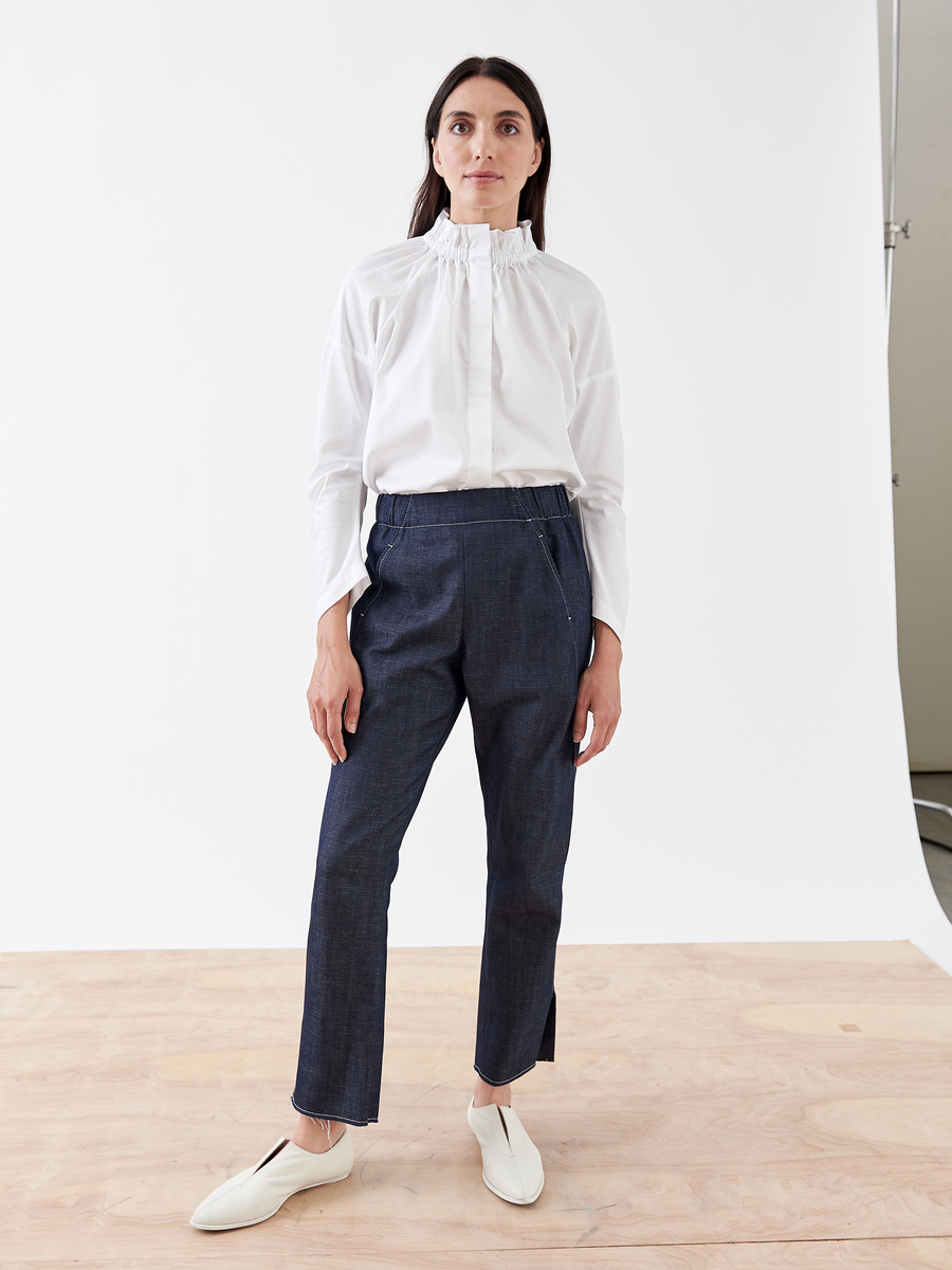 A woman stands on a wooden floor with a white backdrop, dressed in a white ruffled blouse, Zero + Maria Cornejo's Denim Elli Pant - Final Sale, and white slip-on shoes.