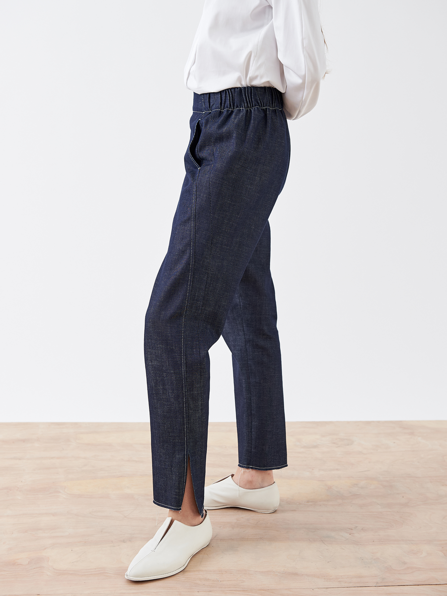 A person in a white long-sleeve top, Denim Elli Pant by Zero + Maria Cornejo with a hem slit, and white shoes stands on a light wood floor. The dark blue elastic-waist pants enhance comfort and style against the plain white background.