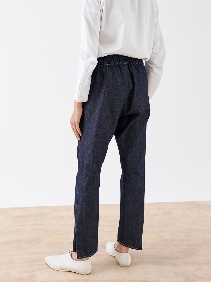 A person wears a white long-sleeve shirt and Denim Elli Pant by Zero + Maria Cornejo, standing on a wooden floor with their back to the camera. They have white shoes, and the background is a plain white wall.