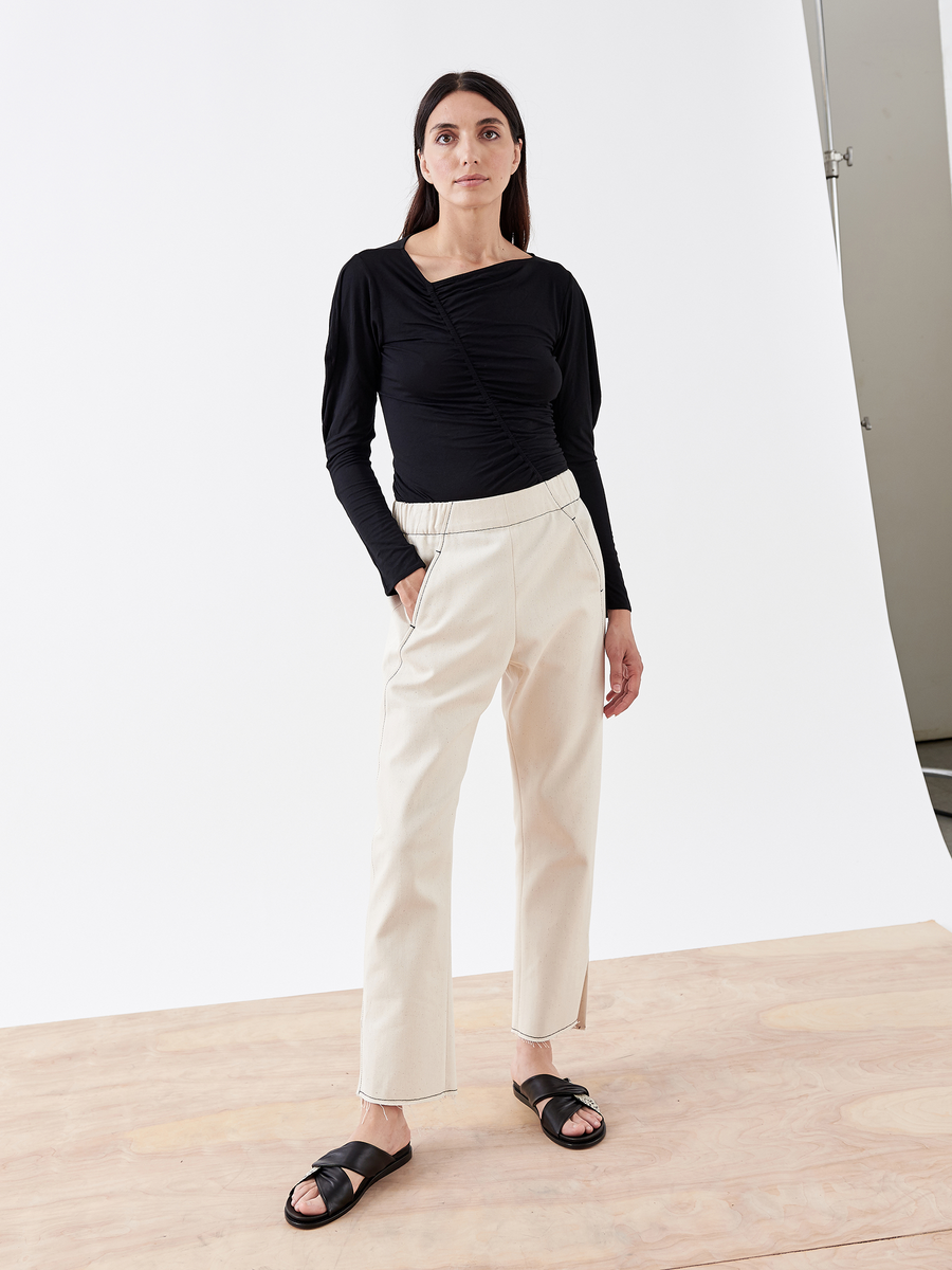 A person with long dark hair poses against a white background in a black, long-sleeve ruched top and Zero + Maria Cornejo's Denim Elli Pant - Final Sale. They complete the look with black sandals.