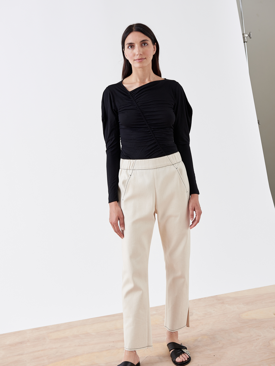 A woman stands against a white background, wearing an asymmetrical black long-sleeve top and the cream-colored Denim Elli Pant by Zero + Maria Cornejo, made from organic denim. With long dark hair and black sandals, she is complemented by the light wood-tone floor in her eco-friendly style.