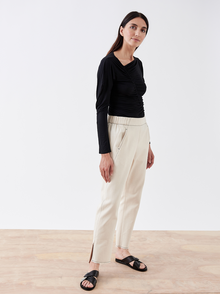 A woman stands confidently on a wooden floor in the Denim Elli Pant by Zero + Maria Cornejo, styled with a black long-sleeve top and black sandals against a plain white background.