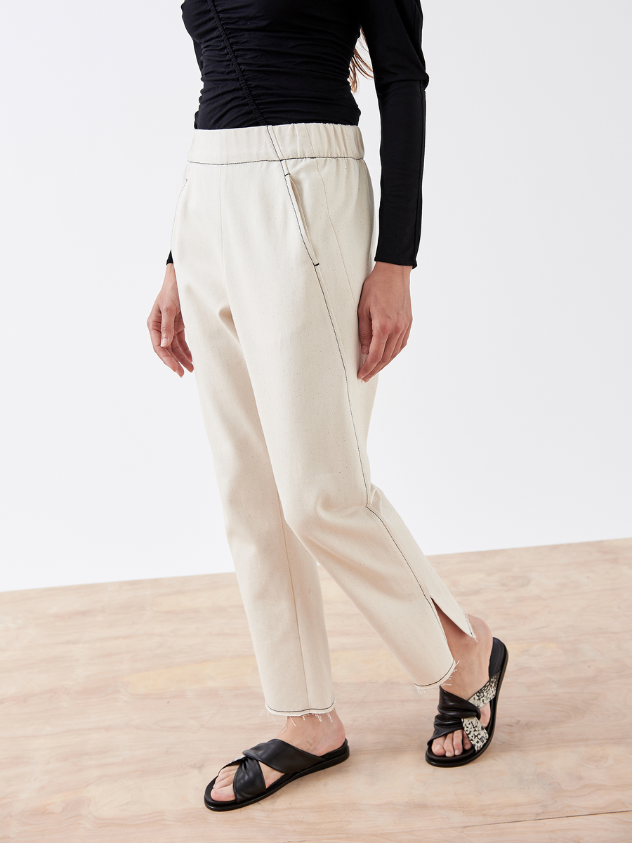 A person is wearing the Zero + Maria Cornejo Denim Elli Pant, off-white with a small slit at the ankle, a black long-sleeve top, and black slide sandals. They stand on a light wooden floor against a plain white background.