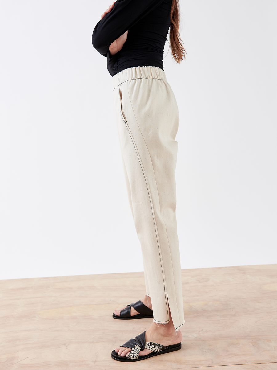 A person in a black long-sleeve top and black slide sandals with a decorative strap crosses their arms, wearing Zero + Maria Cornejo's Denim Elli Pant in organic cotton beige. The background is plain.