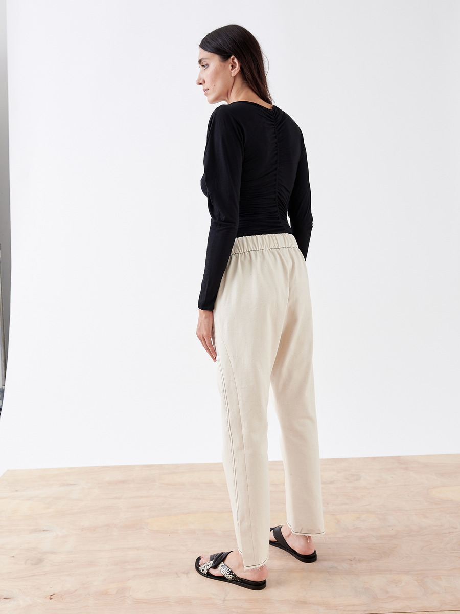 On a wooden floor, a person with long dark hair and a plain white wall backdrop wears a black long-sleeve top, Denim Elli Pant by Zero + Maria Cornejo, and black sandals.