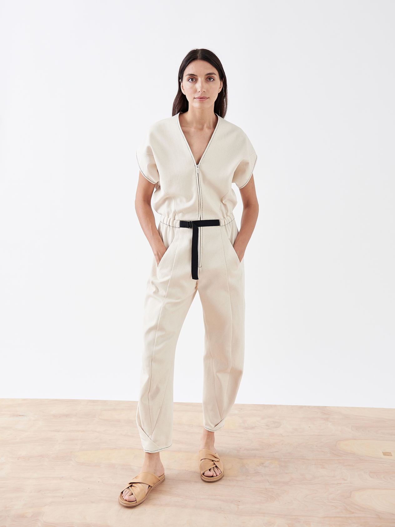A person with long brown hair stands against a plain white wall, wearing the dark Strap Akeo Jumpsuit made of 100% organic cotton denim from Zero + Maria Cornejo. The jumpsuit features short sleeves, a zippered front, and is belted at the waist. They have their hands in their pockets and are wearing black slip-on shoes.