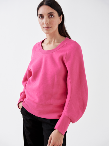 A person with long dark hair wears the B Sweatshirt by Zero + Maria Cornejo, featuring a bright pink color and puffed sleeves in sustainable fashion, paired with black pants. They stand against a plain white background, looking at the camera.