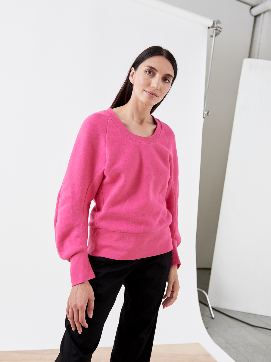 A person with long dark hair wears the B Sweatshirt - Final Sale from Zero + Maria Cornejo in bright pink biodegradable fabric and black pants. They stand in a studio with a white backdrop, facing the camera, embodying sustainable fashion.