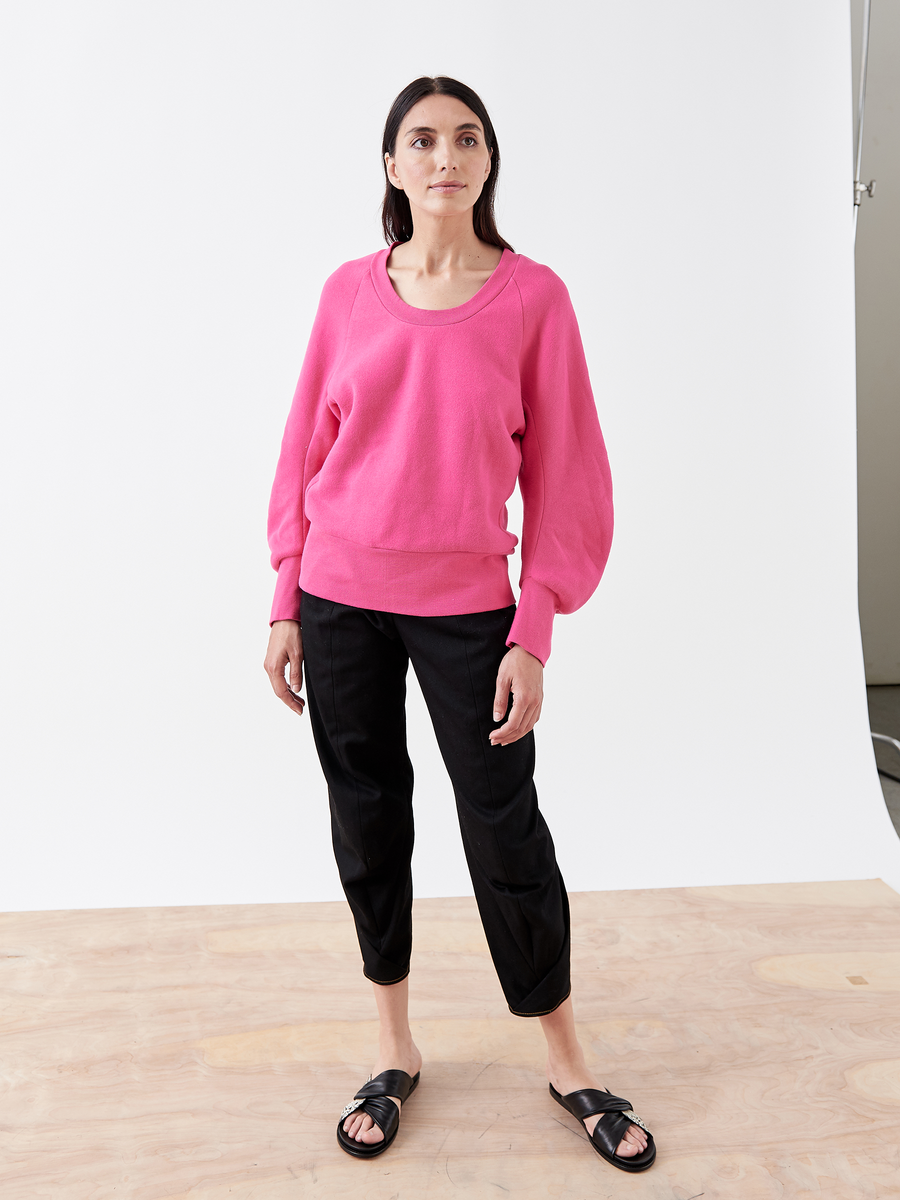 A person with long dark hair stands on a wooden floor against a white backdrop, wearing the B Sweatshirt by Zero + Maria Cornejo in bright pink biodegradable fabric, black pants, and black sandals. Their hands rest at their sides as they look forward calmly.