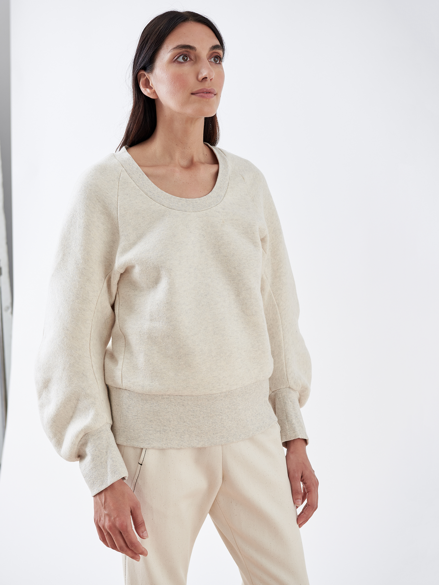 A person with long dark hair wearing the B Sweatshirt by Zero + Maria Cornejo, crafted from biodegradable fabric in light beige, and matching pants stands against a plain white background, looking slightly upward to embody sustainable fashion.