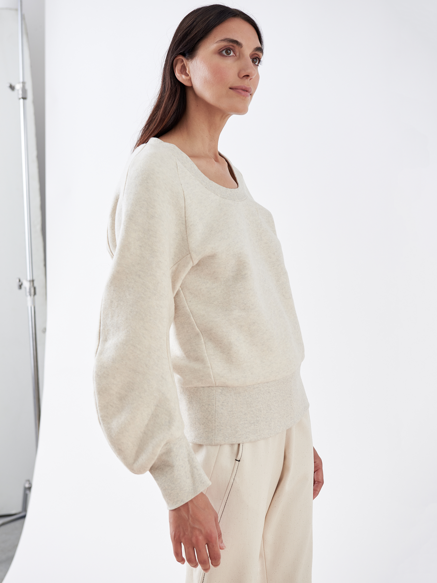 In a studio, a woman models the B Sweatshirt from Zero + Maria Cornejo. She is dressed in light beige biodegradable fabric and gazes upwards with her hands at her sides against a plain white backdrop with a metal stand to her left, capturing sustainable fashion perfectly.