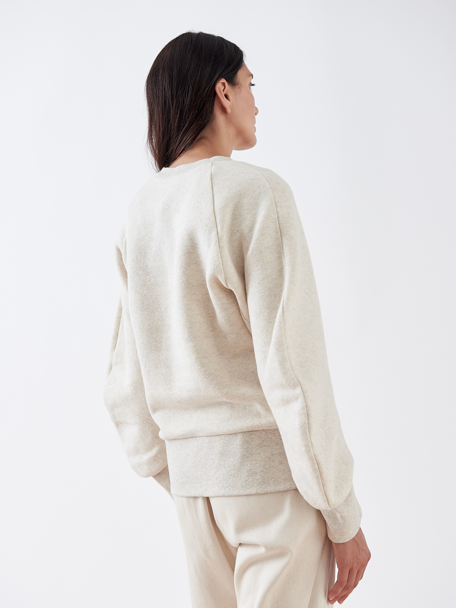 A person with long dark hair is wearing a light gray B Sweatshirt - Final Sale from Zero + Maria Cornejo and cream-colored, biodegradable pants, standing against a plain white background. They are slightly turned to their left, with their face not visible.