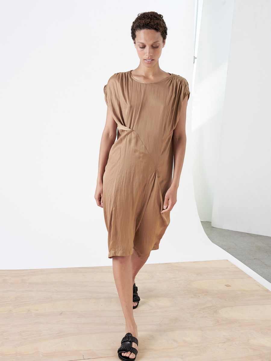 A woman in the Ruched Fold Dress by Zero + Maria Cornejo, featuring a tan washed georgette with an asymmetrical front, strolls across a light wooden floor in black sandals. The minimalist setting has white walls and reveals an arched, light-filled area.