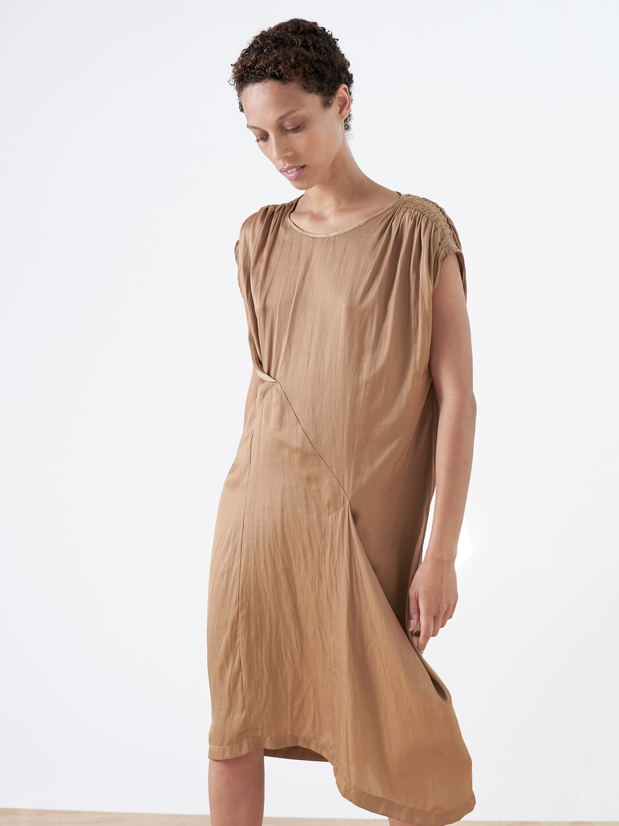 Someone with short curly hair stands against a white background, wearing the Ruched Fold Dress - Final Sale by Zero + Maria Cornejo. The dress is light brown washed georgette with an asymmetrical folded front and hemline, creating a relaxed posture as they look down.