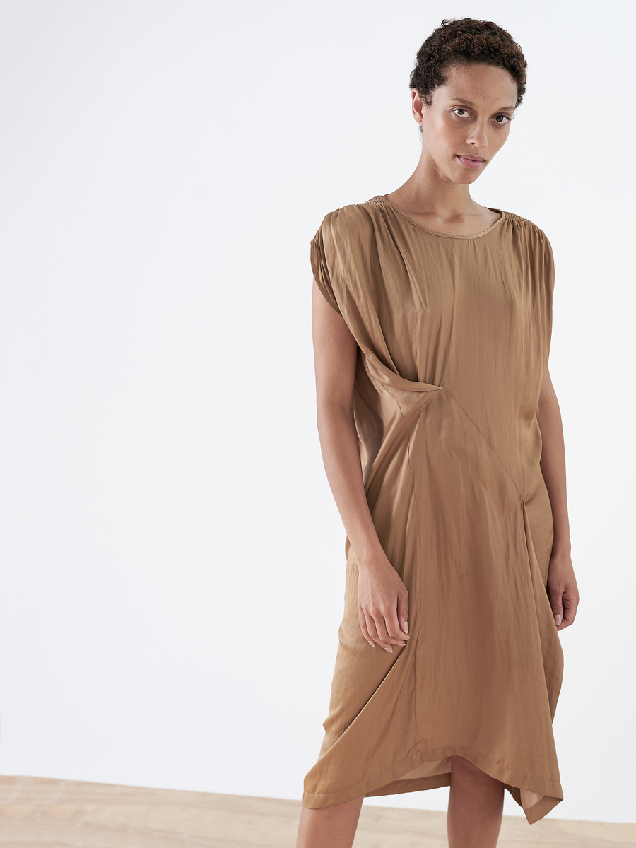 A person models the Ruched Fold Dress - Final Sale by Zero + Maria Cornejo in a minimalist setting. This loose, asymmetrical brown dress with washed georgette fabric has gathered shoulders and side, forming soft drapes against a light floor and plain white wall.