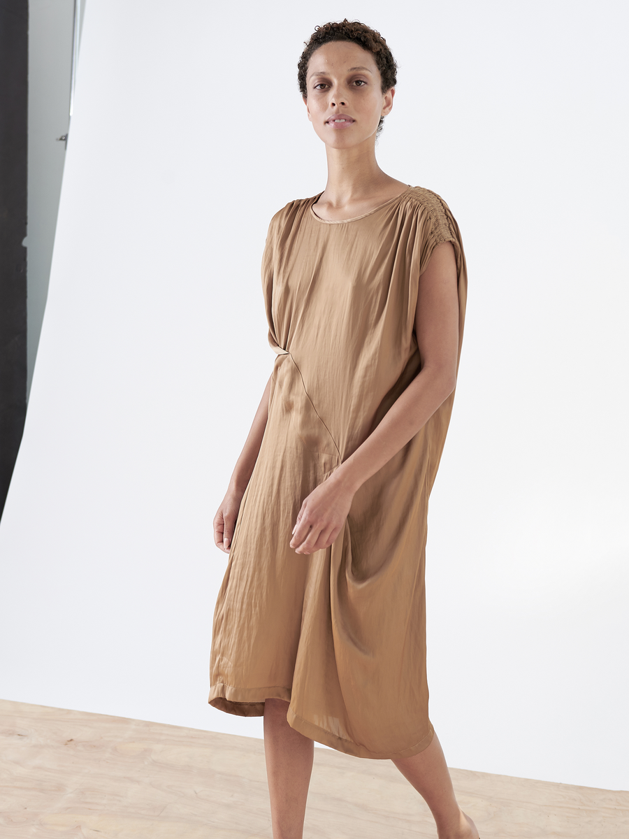 A person with short curly hair wears the Ruched Fold Dress by Zero + Maria Cornejo, a light brown asymmetrical piece in washable fabric. They walk on a wooden floor against a neutral white backdrop.