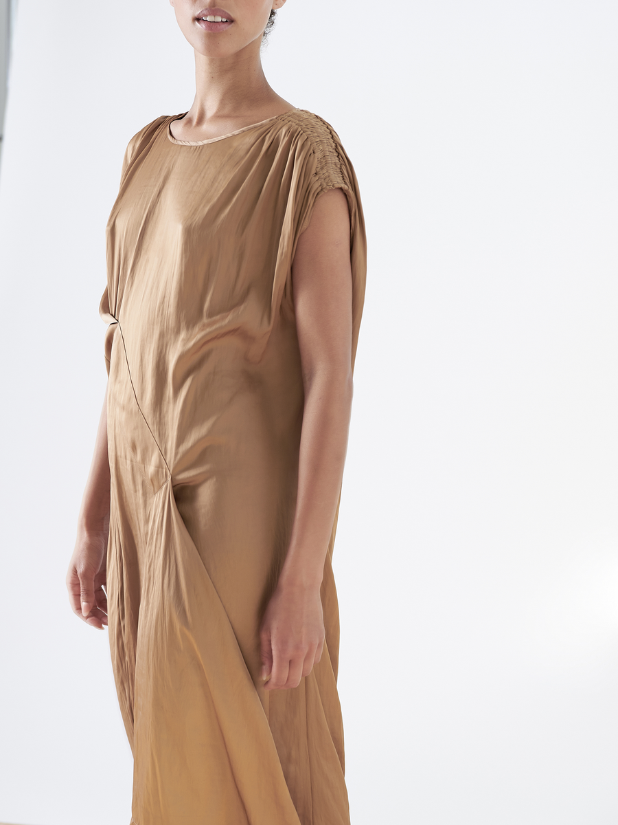 A person is wearing the Ruched Fold Dress by Zero + Maria Cornejo, a tan washed georgette dress with a subtle sheen. It has short sleeves, gathered shoulder details, and an asymmetrical folded front. The white background enhances its minimalist elegance.