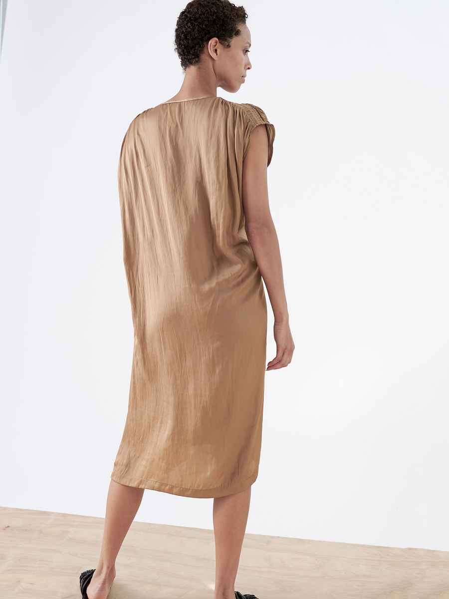 On a light wood floor, someone models the Ruched Fold Dress by Zero + Maria Cornejo, a sleeveless ochre dress made from washable fabric. The asymmetrical folded front adds elegance as they turn, showcasing the dress's back and side against a plain white backdrop.