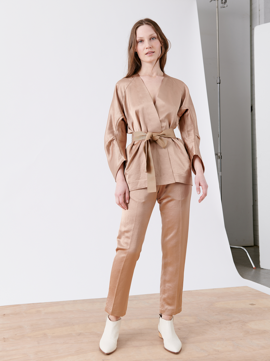 A woman models the Curve Oki Jacket from Zero + Maria Cornejo, a light brown satin cardigan-style jacket with a grosgrain tie belt, paired with matching pants. She stands neutrally on a wooden floor against a white backdrop, wearing chic white ankle boots.