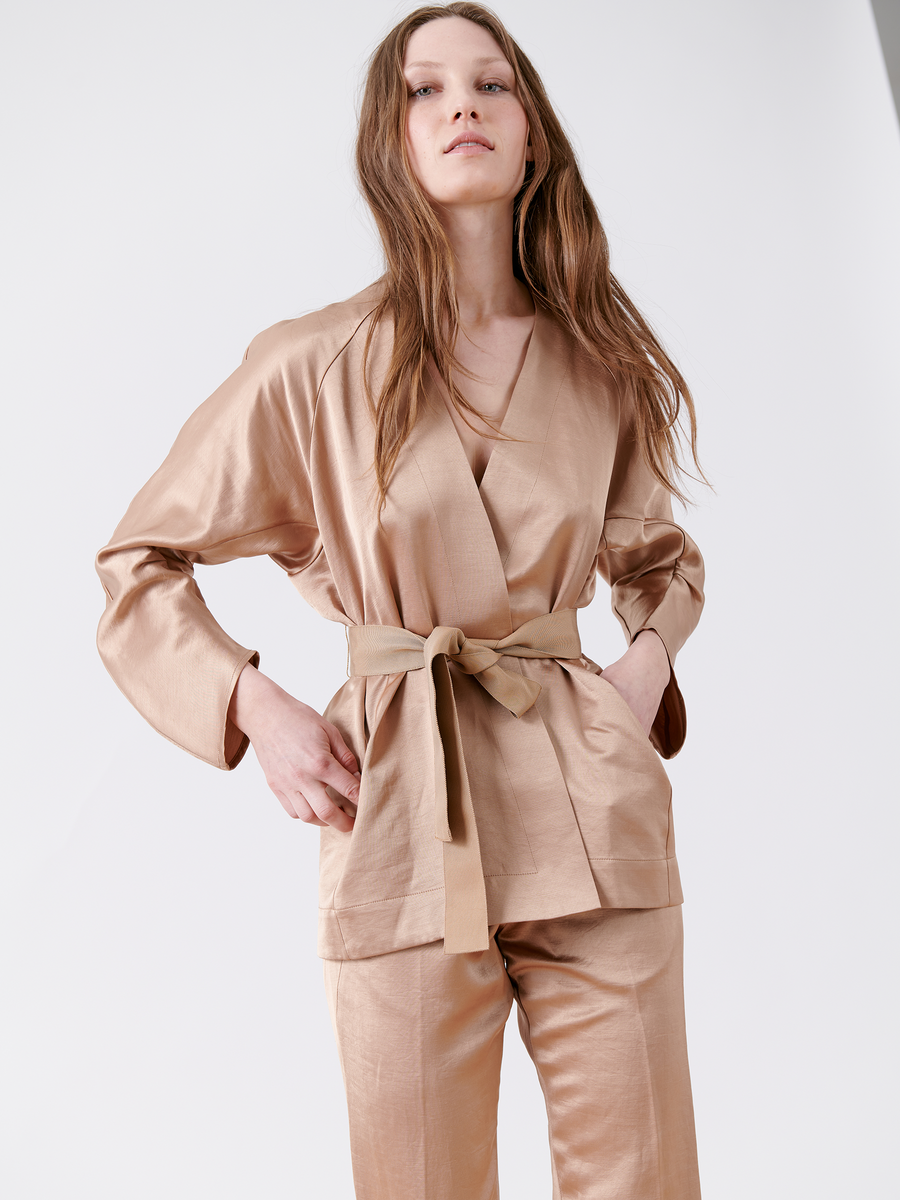 A person with long brown hair stands confidently in a sophisticated outfit featuring the Curve Oki Jacket by Zero + Maria Cornejo, paired with a silky beige wrap top, grosgrain tie belt, and matching pants. The ensemble is set against a simple light gray background.