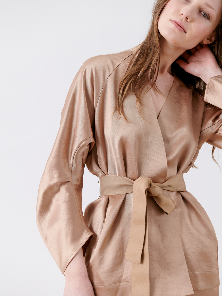 A woman in a beige Curve Oki Jacket by Zero + Maria Cornejo, with wide sleeves and a grosgrain tie belt, tilts her head slightly and touches her neck. Her long hair contrasts with the plain white background, highlighting the attire's elegance reminiscent of a satin wrap dress.