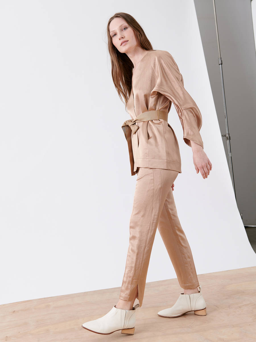 A long-haired woman models the Curve Oki Jacket from Zero + Maria Cornejo, paired with matching linen pants and white ankle boots. She poses on a wooden floor against a white studio backdrop, looking sideways.