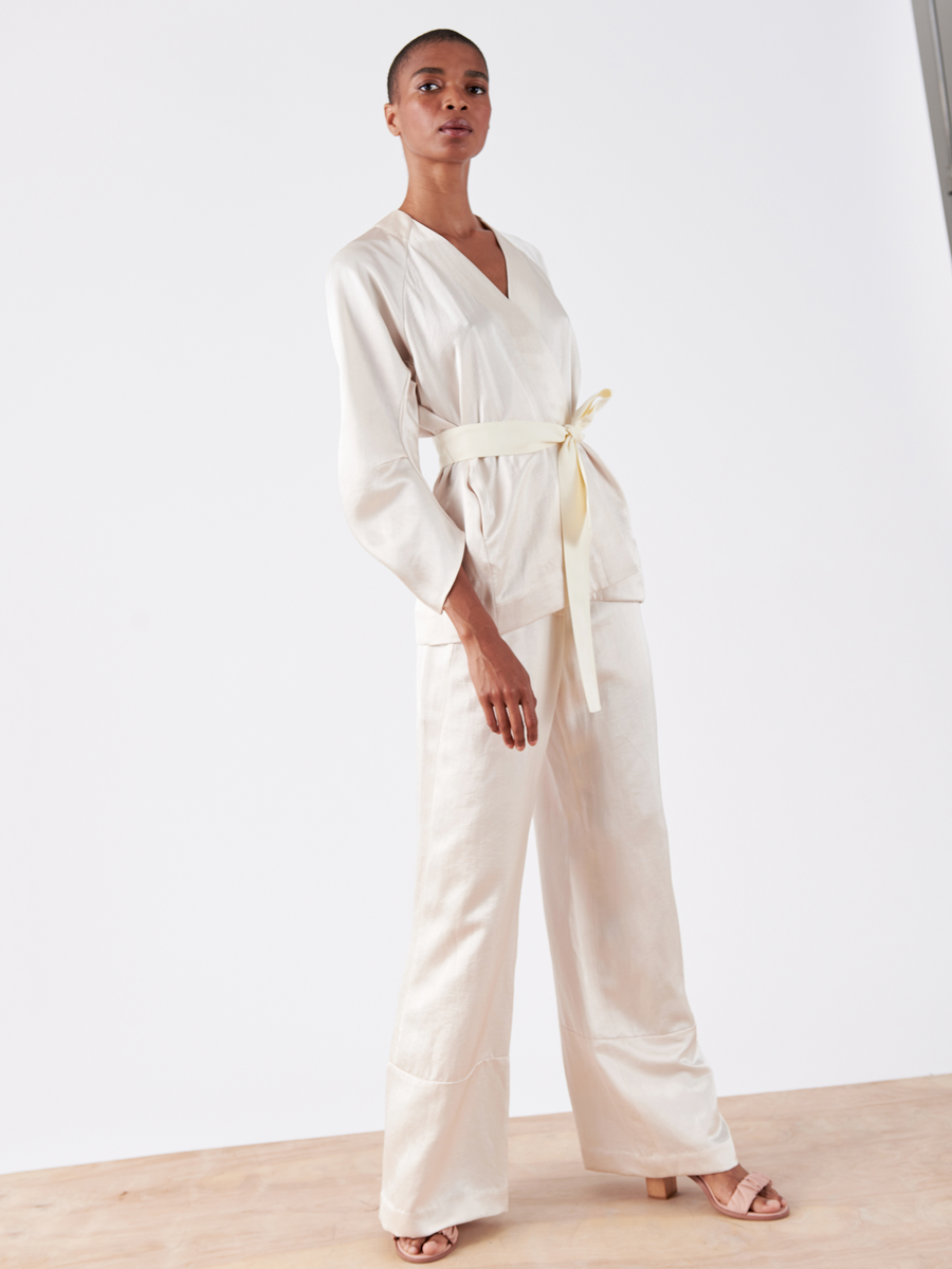 A person confidently models the Curve Oki Jacket by Zero + Maria Cornejo, a cream-colored, cardigan-style jacket with a grosgrain tie belt and wide-legged linen-blend trousers, boasting a silky texture. The ensemble is set on a wooden floor against a plain white background.