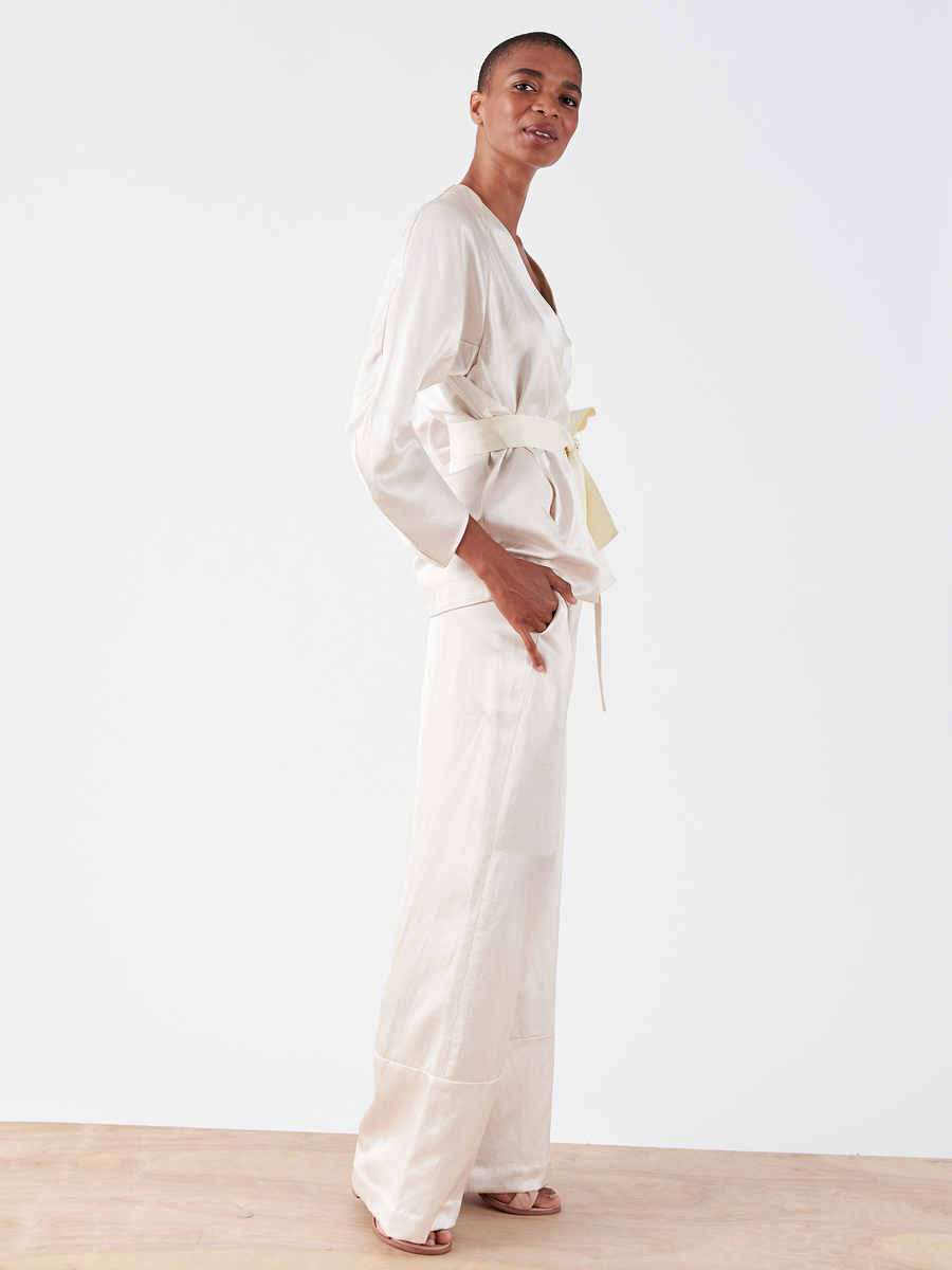 Wearing the Curve Oki Jacket from Zero + Maria Cornejo, a person stands sideways on a light wood floor. The light-colored ensemble, featuring a grosgrain tie belt and wide-leg pants, showcases elegant simplicity and craftsmanship against a plain white background.