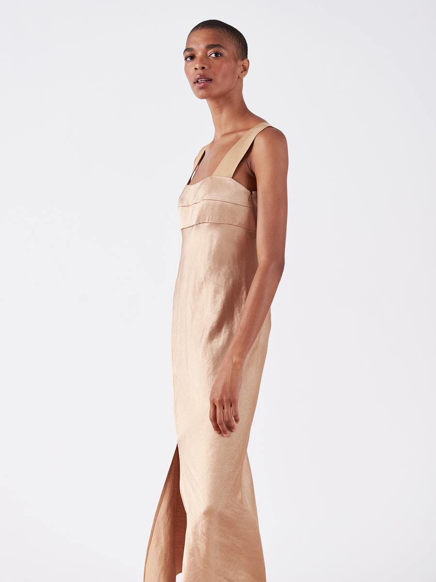 A person is wearing the Bias Oona Dress - Final Sale by Zero + Maria Cornejo, featuring a sleeveless, floor-length satin design in beige with a square neckline and a slit. They're standing against a plain white background, looking slightly to the left.