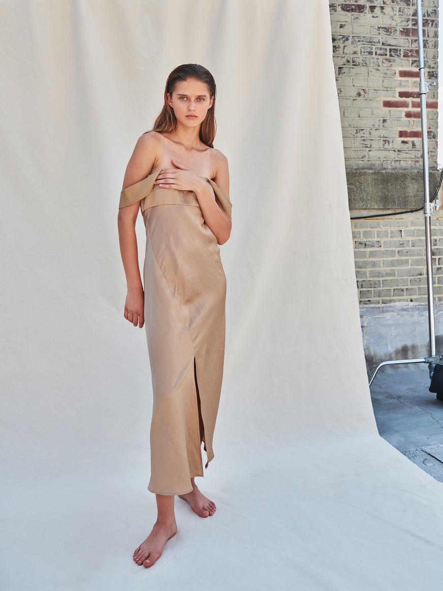 A barefoot woman on a studio set models the Zero + Maria Cornejo Bias Oona Dress - Final Sale, showcasing its elegant silhouette with one hand on her chest. She stands before a white cloth and brick wall.