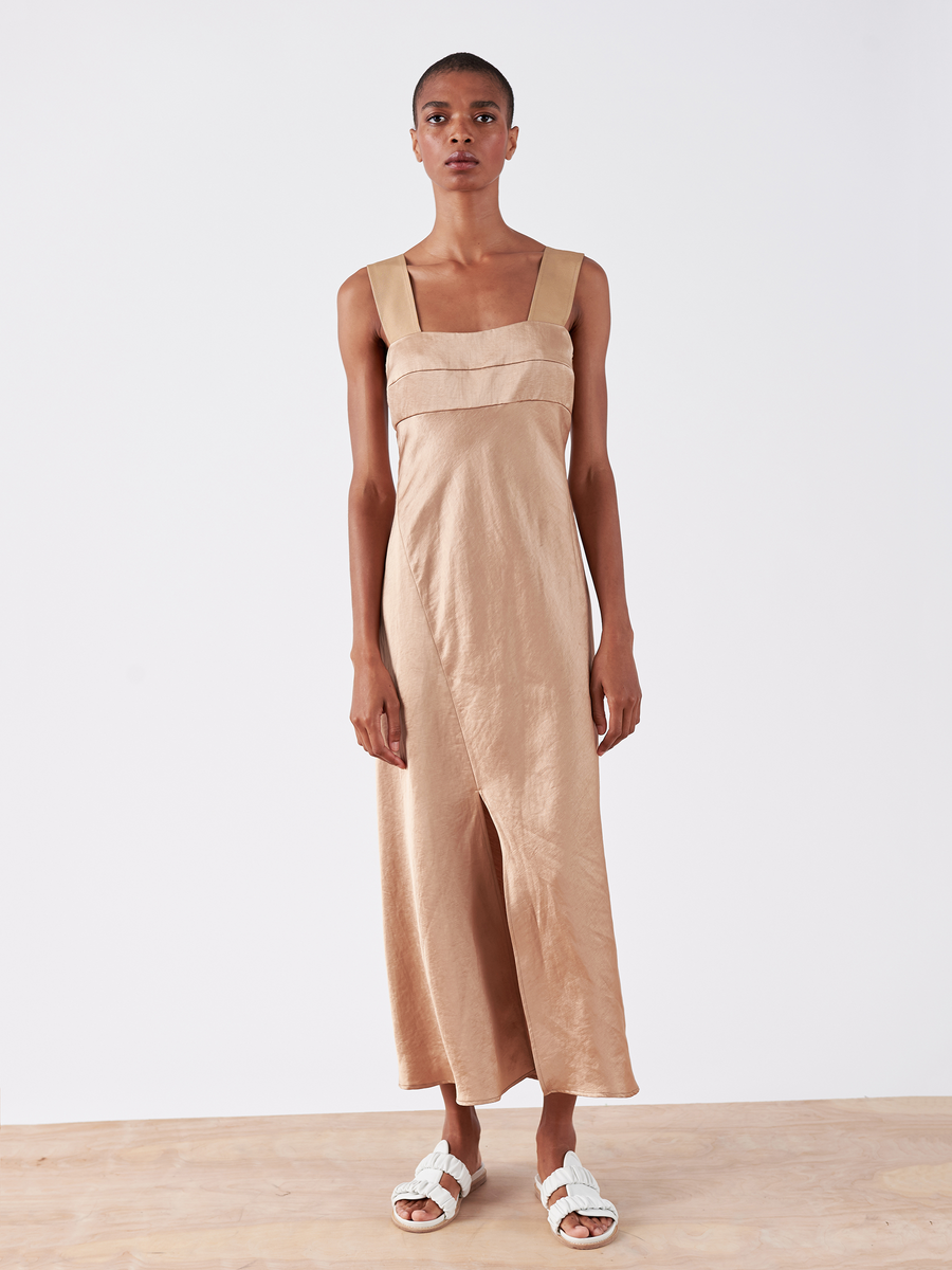 A person stands in the Bias Oona Dress from Zero + Maria Cornejo, featuring wide straps, a square neckline, and a bias silhouette with a front slit. The beige linen-blend fabric has a slight sheen. They complete the look with white sandals on a wooden floor against a simple backdrop.