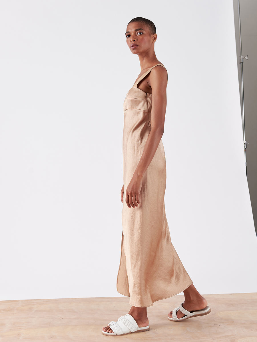 A person wearing the Bias Oona Dress - Final Sale by Zero + Maria Cornejo, paired with white sandals, stands on a wooden floor against a plain white background, looking to the side.